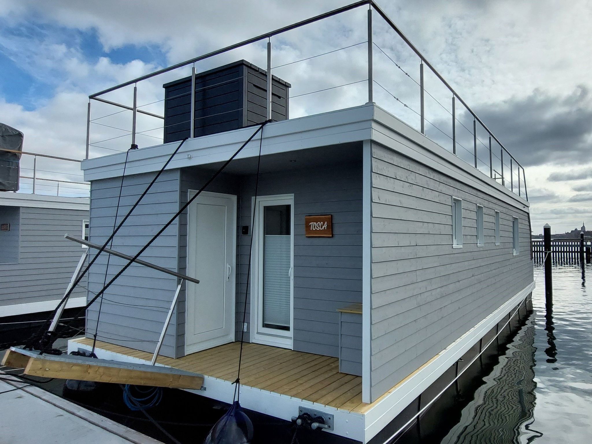 Une maison flottante moderne dotée d’une terrasse, d’une porte portant l’inscription « TOSCA » et offrant une vue sur l’eau.
