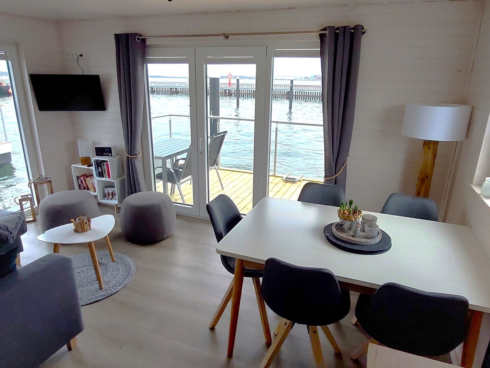 A modern living room with a white table, grey sofa, and a balcony with a view of the water.