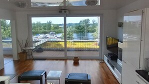 Ruime kamer met een grote schuifpui naar een balkon met uitzicht op een meer en boten. Aan de rechterkant staat een televisie en in het midden staat een klein tafeltje met stoelen.