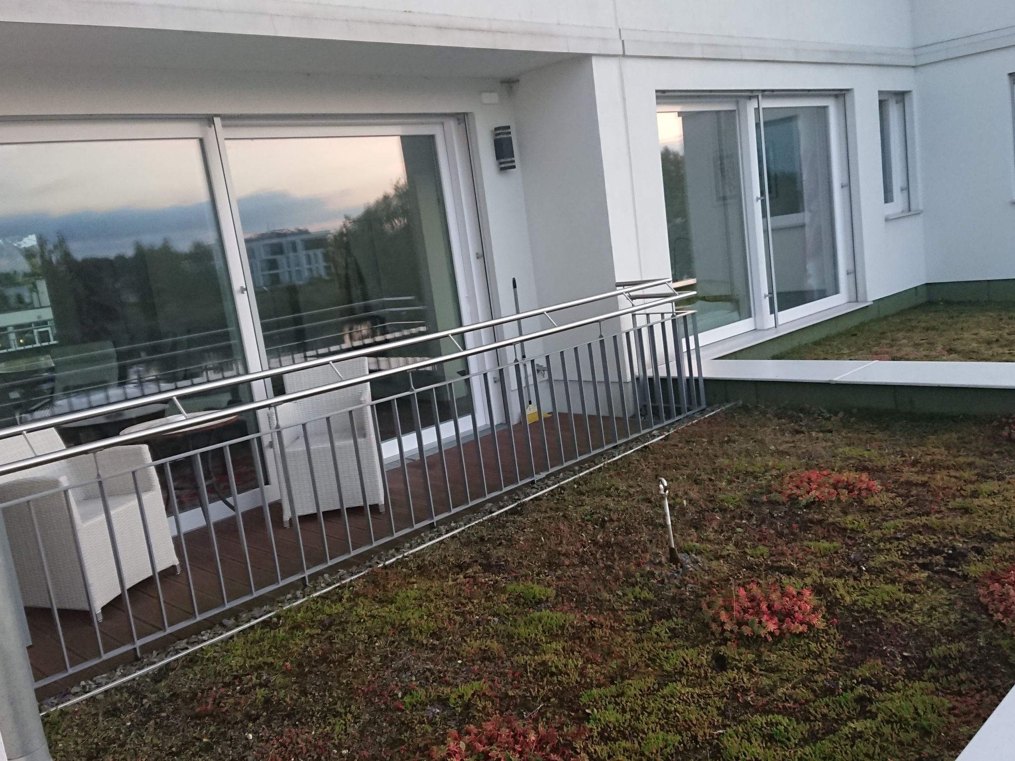 En balkon med gelænder, et lille haveareal med røde blade og udsigt til en bygning gennem store vinduer.