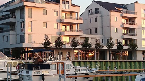 Een meerlaags woongebouw met balkons, een steiger met boten en een groene wand.