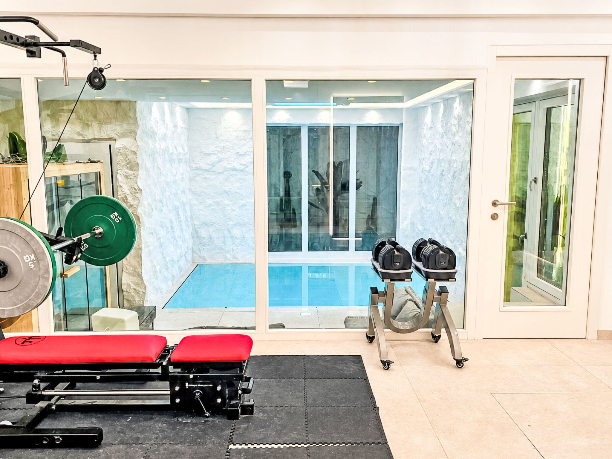 Ein Fitnessraum mit Krafttrainingsgeräten, einer Bank und einem Schwimmbereich, der durch Glastüren einsehbar ist.
