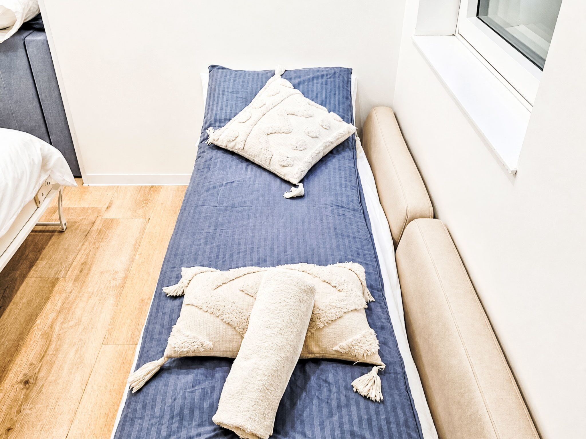 Ein Einzelbett mit blauer Matratze und weißen Kissen, ein Holzboden und ein Fenster mit weißen Jalousien.
