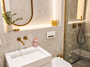 Ein modernes Badezimmer mit einem weißen Waschbecken, goldenen Armaturen, einer Topfpflanze und einer Duschkabine aus Glas.