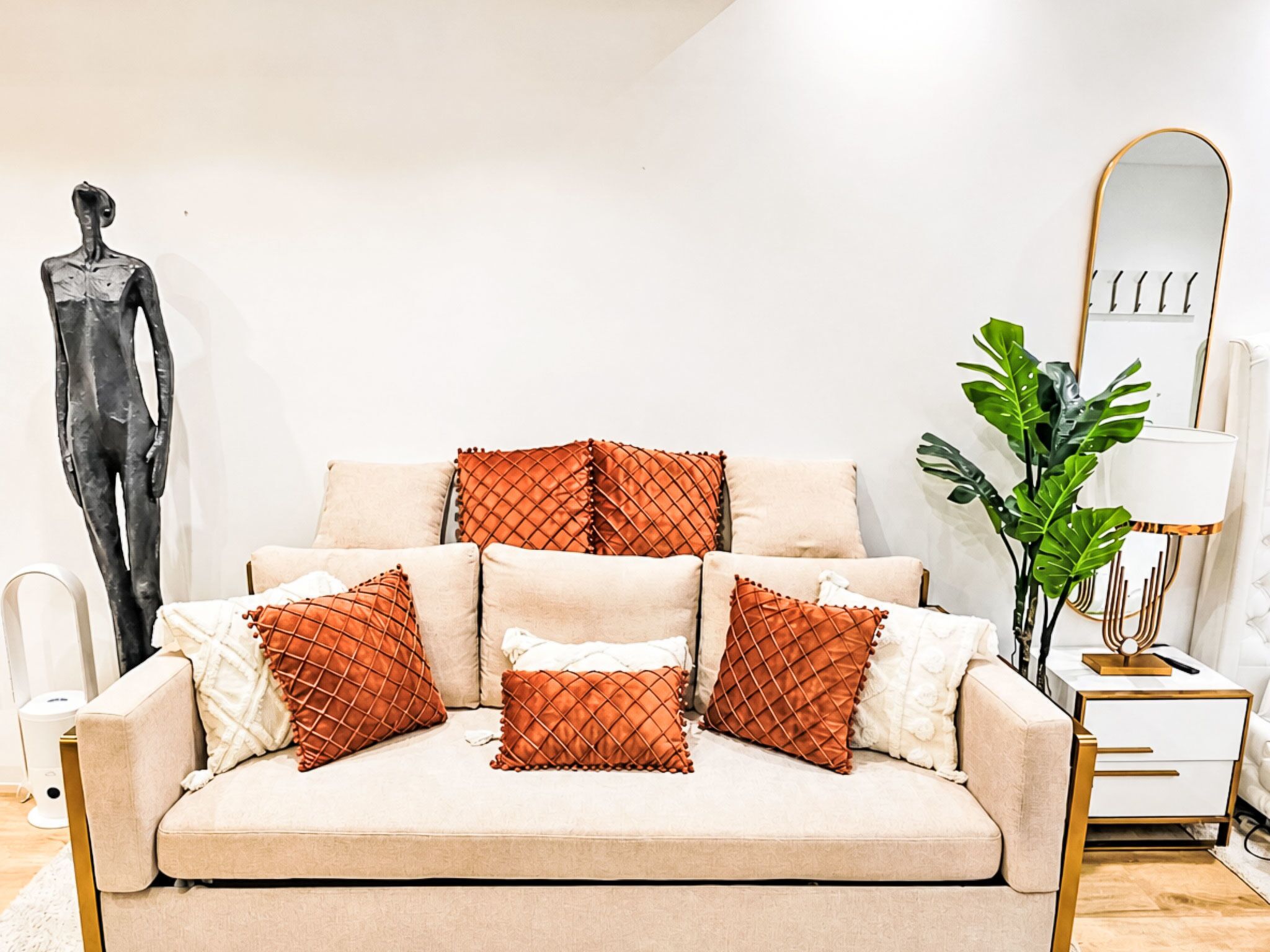 Ein beiges Sofa mit gemusterten Kissen, eine minimalistische Skulptur, eine moderne Lampe und eine große Pflanze.