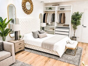 Een slaapkamer met een gestoffeerd hoofdeinde, een nachtkastje met een lamp, een grote spiegel, een kledingkast met hangende kleding en een potplant.
