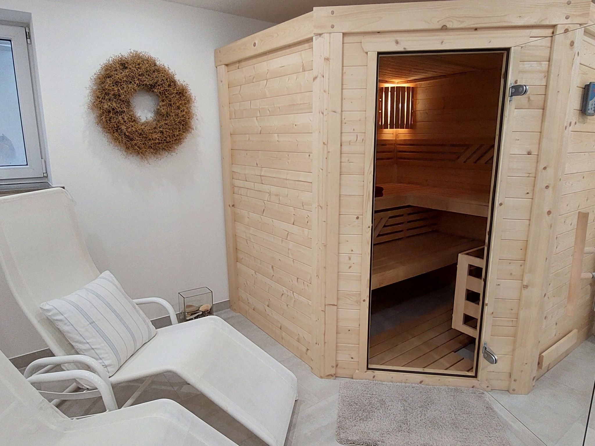 Una sauna con rivestimento in legno, una porta e un piccolo gradino d'ingresso. Si vede una sedia bianca e un cuscino bianco all'esterno.