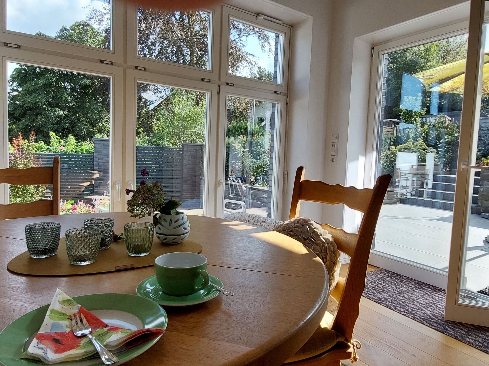 A dining area with a round table, chairs, and a view of a garden through large windows.