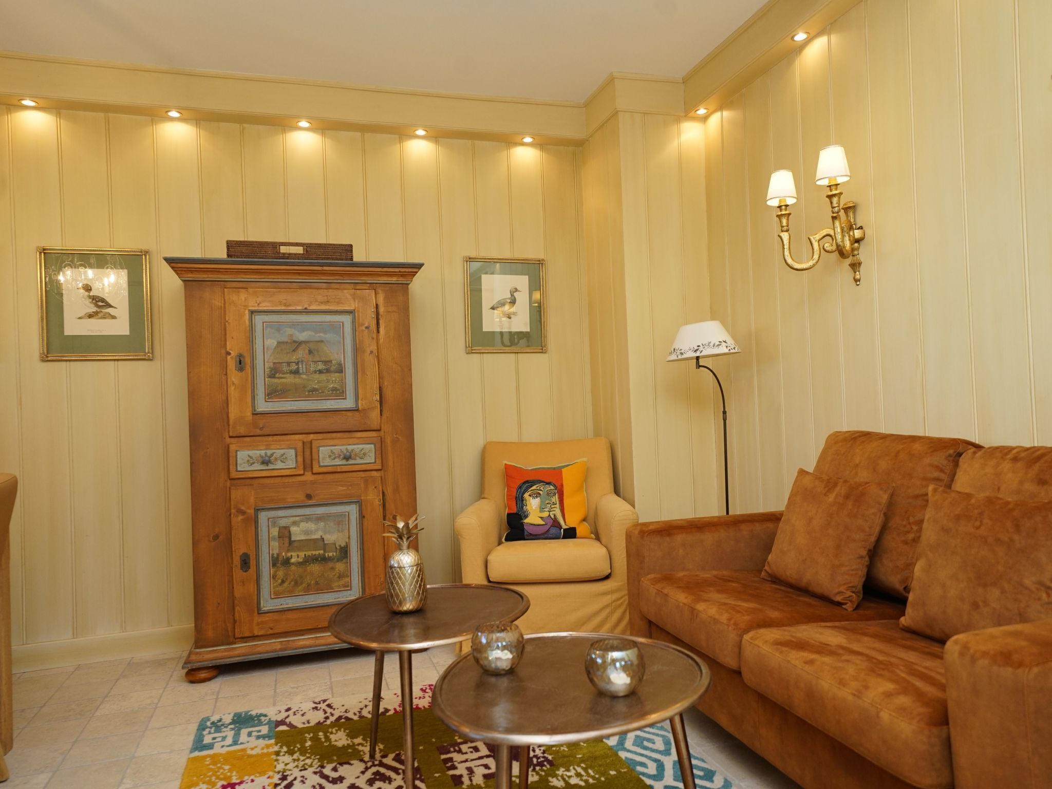 Una sala de estar acogedora con un sofá marrón, un mueble de madera con cuadros enmarcados, una mesita redonda y una lámpara de pie.