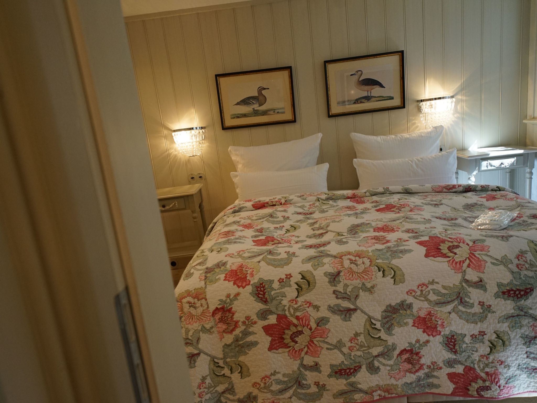 A hotel room with a floral bedspread, two framed wall art pieces, a nightstand with a lamp, and a bedside table with a lamp.