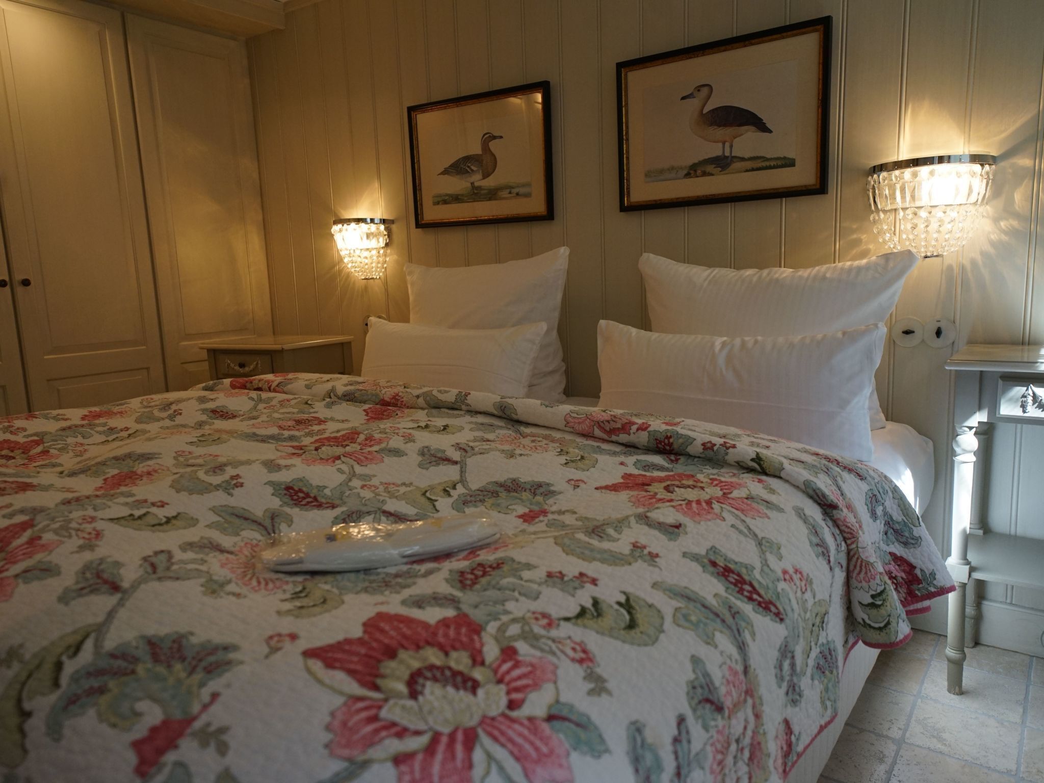 A bedroom with a floral bedspread, two framed wall art pieces, a nightstand with a lamp, and a white door.