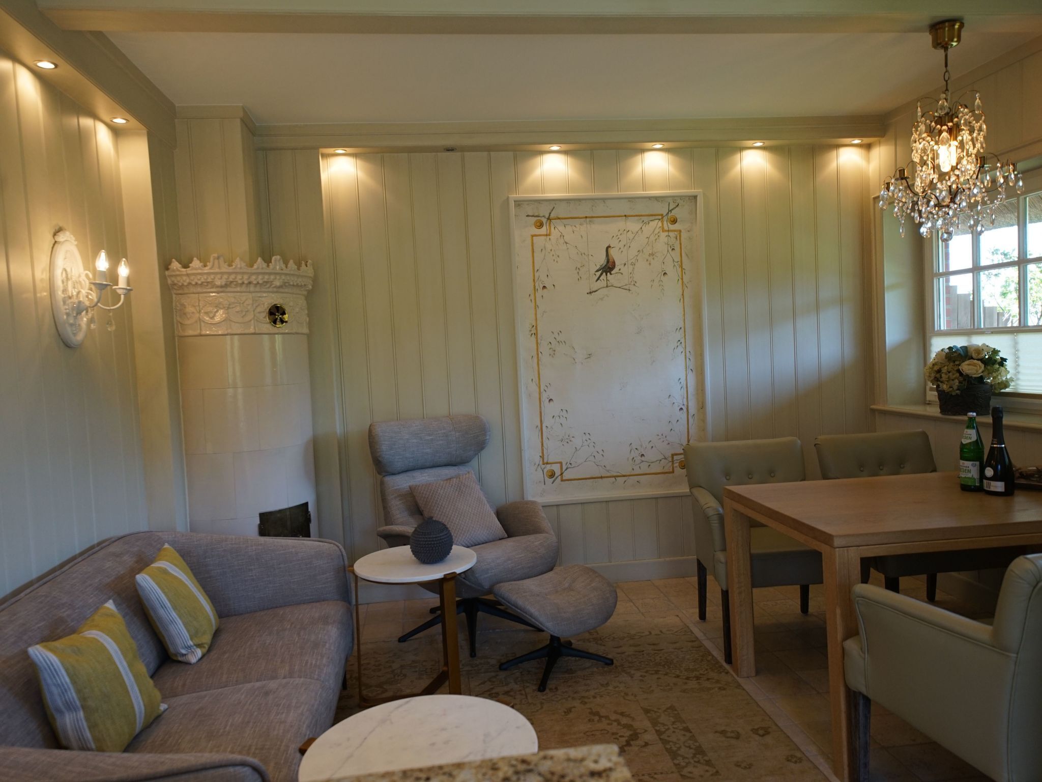 A cozy living room with a sofa, armchair, and a small table. There is a decorative wall piece and a chandelier.