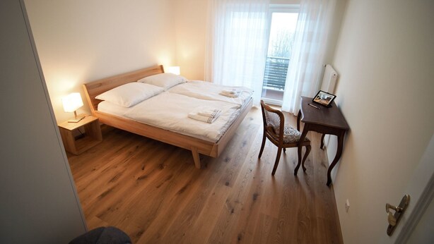 A double bed with two pillows, a bedside table with a lamp, a wooden chair, a small desk with a picture frame, and a window with sheer curtains. - Haus an der Lieser by Interhome (Spittal an der Drau)