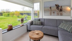 Een moderne woonkamer met een grijze bank, een ronde houten salontafel en een groot raam met uitzicht op een groene tuin.