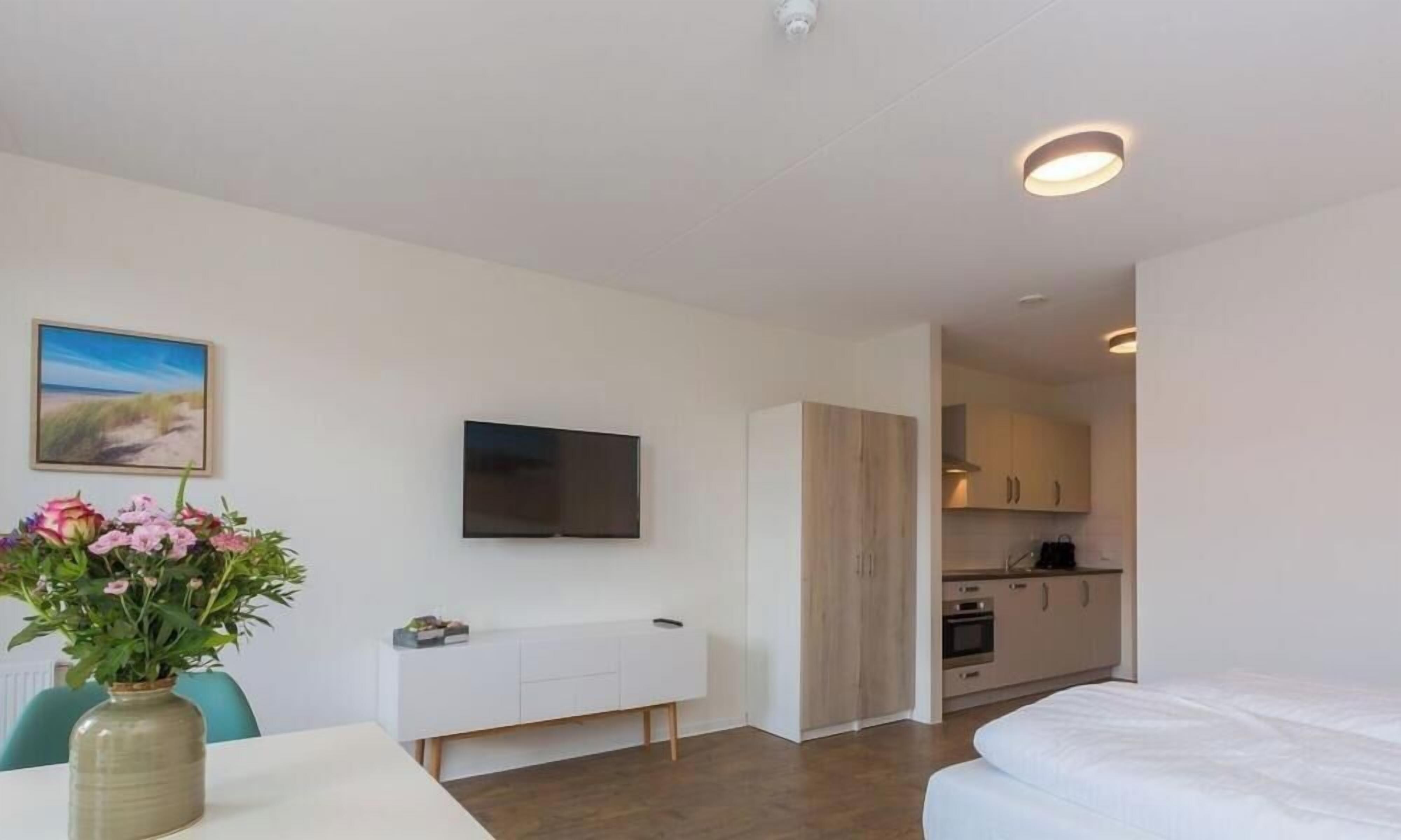 Une chambre d’hôtel moderne dotée d’une télévision à écran plat, d’un coin repas agrémenté d’un vase de fleurs et d’une kitchenette.