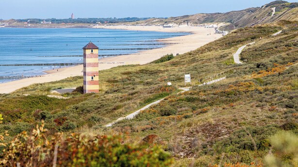 Paisagem costeira com farol, praia e terreno montanhoso.