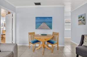 A dining area with a round wooden table and two chairs, a painting of a pier on the wall, and a view of the ocean through the window.