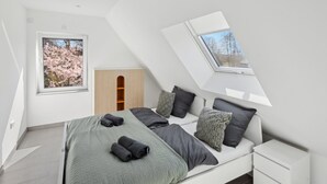 Ett modernt sovrum med ett sluttande tak, ett takfönster, en säng med grå sängkläder och en inramad tavla med körsbärsblommor.