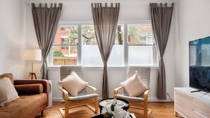 A living room with a brown sofa, two armchairs, a coffee table with a vase of flowers, a TV, and a window with curtains.