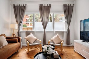 Un salón con un sofá marrón, dos sillones, una mesa de centro con un jarrón de flores, un televisor y una ventana con cortinas.