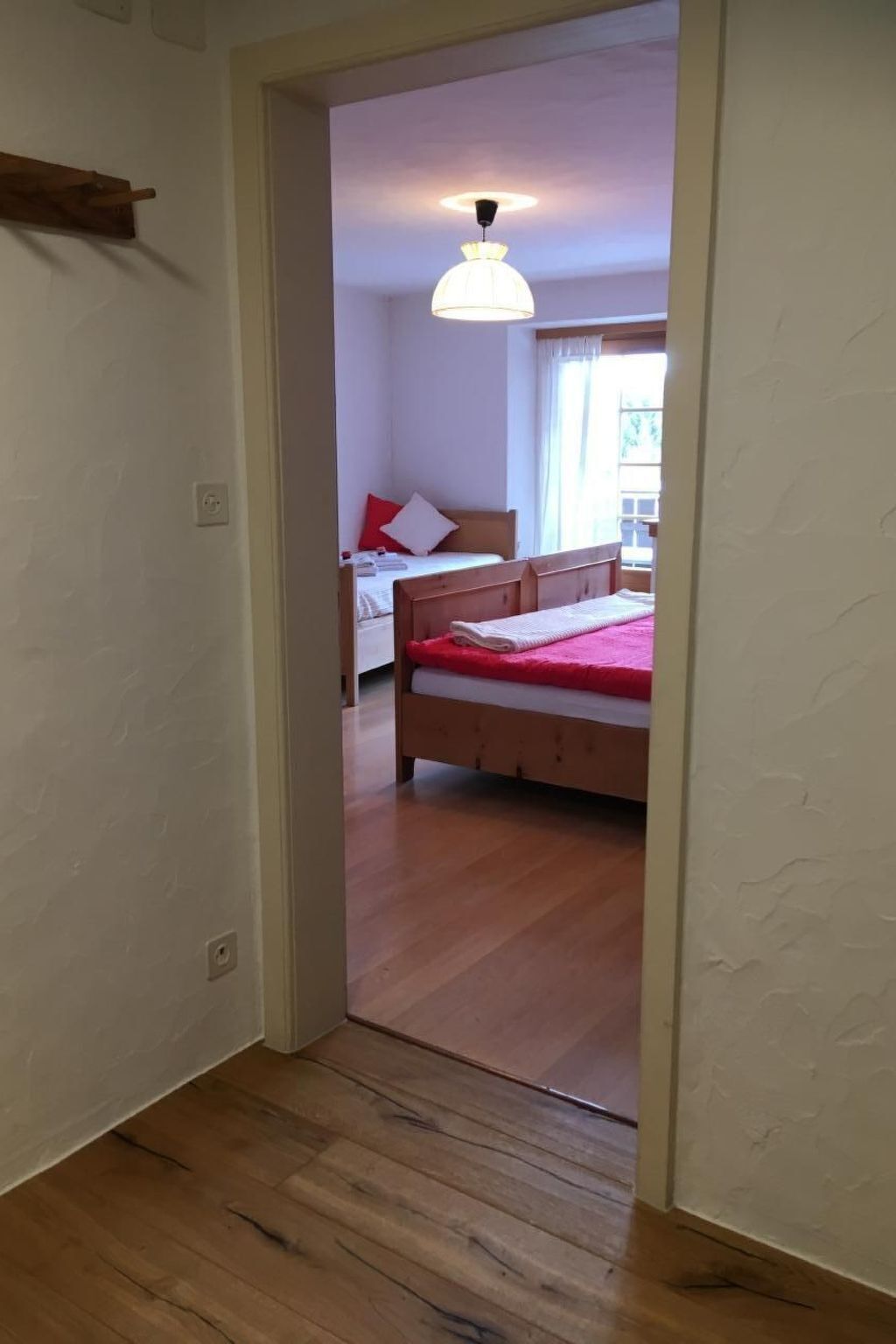 A narrow hallway leading to a bedroom with a wooden floor, a bed with a red blanket, and a white ceiling light.
