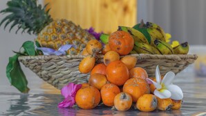Una cesta de mimbre llena de diversas frutas, como naranjas, plátanos y una piña, sobre una superficie reflectante.