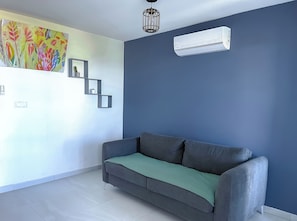 A modern living room with a grey sofa, a colorful abstract painting, a cactus, and a geometric shelf unit. - Studio Paille en ké close to all amenities (C.H.U, IUT, BEACH) (Saint pierre)