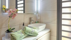 Baño con lavamanos blanco, un espejo y una ventana con persianas. Hay una toalla con estampado de pájaro y una pequeña planta en maceta sobre el mostrador.