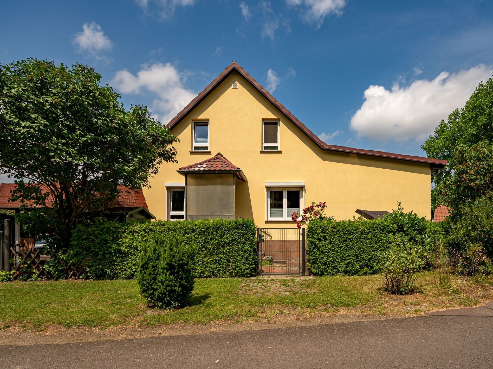 A yellow two-story house with a brown roof, surrounded by greenery and a metal gate.