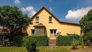 Une maison jaune à deux étages, avec un toit brun, entourée de verdure et fermée par un portail en métal.