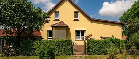 A yellow two-story house with a brown roof, surrounded by greenery and a metal gate.