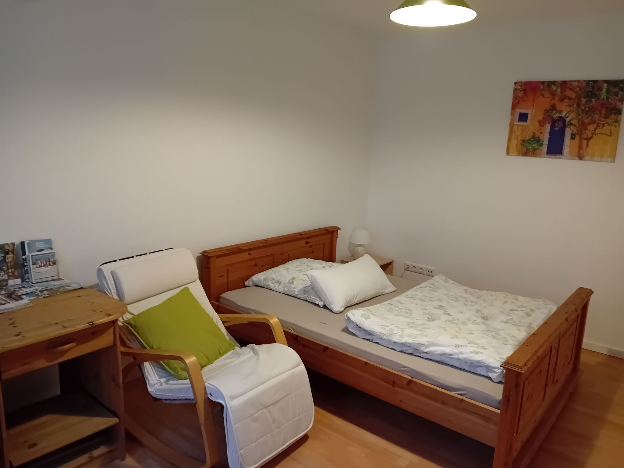 Una habitación pequeña y acogedora con una cama de madera, una silla con un cojín verde, un escritorio con una lámpara y un cuadro en la pared.