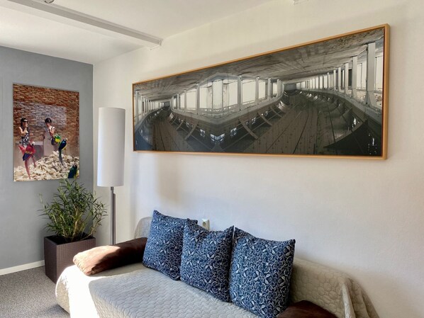 A modern hotel room with a large bed, a potted plant, a wall-mounted painting, and a large window.
