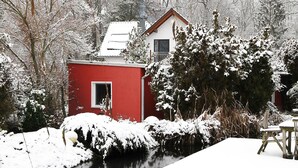 Een rood gebouw met een klein raam, omgeven door met sneeuw bedekte bomen en een bevroren rivier.
