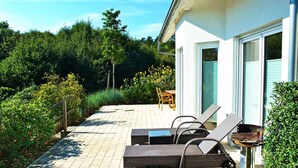Eine Außenpatio mit Liegestühlen und Grill, umgeben von Grünpflanzen und einem Gebäude mit großen Fenstern.