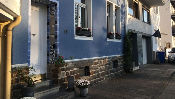 Ein blaues Gebäude mit steinernen Basis, Hausnummer 73, mit einer Tür, Fenstern mit Blumenkästen und einer kleinen Gartenanlage mit Topfpflanzen.