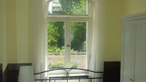 Ein Zimmer mit einem Bett, einem kleinen Tisch, einer Lampe, einem Kleiderschrank und einem Fenster mit Vorhängen.