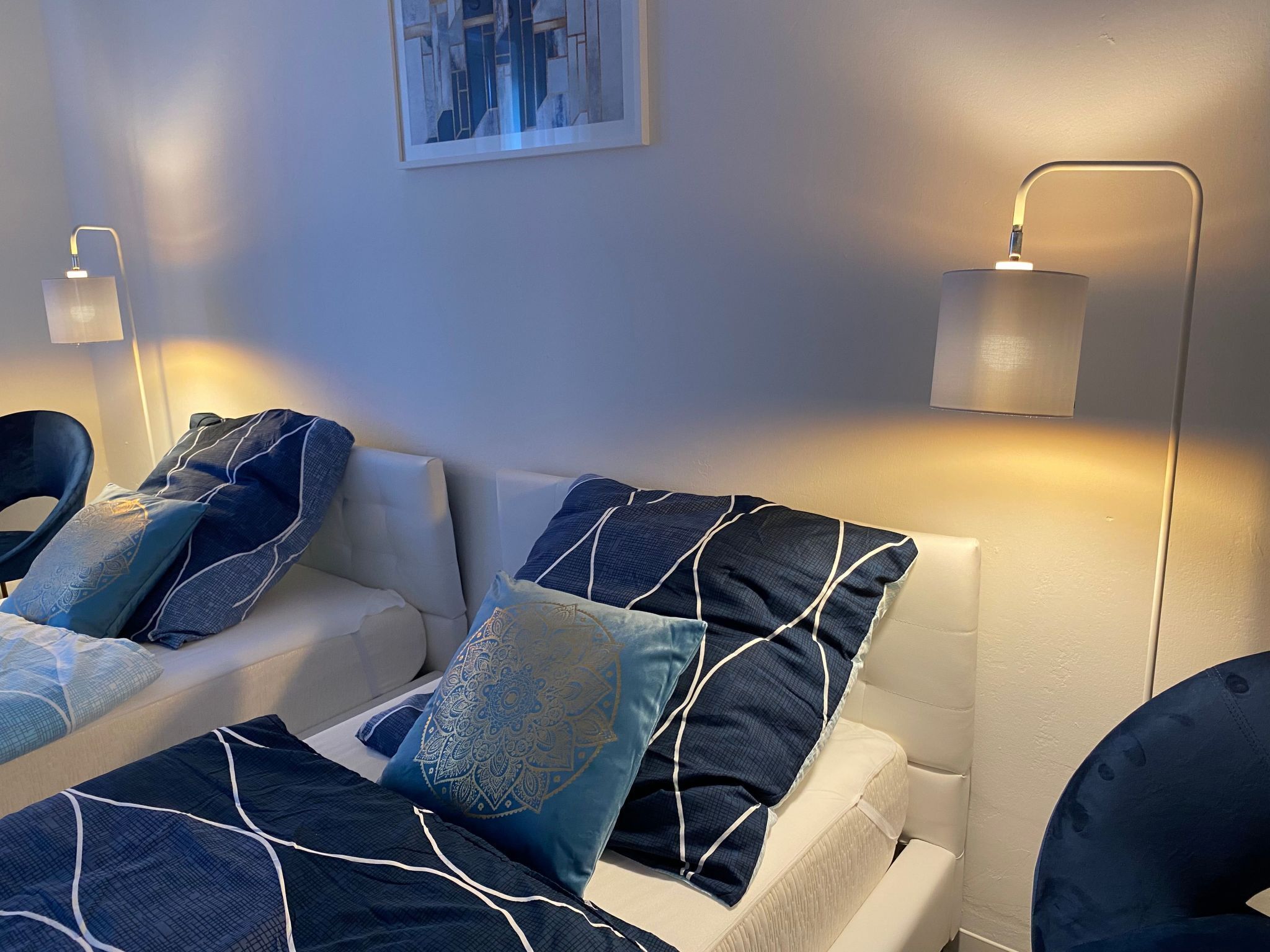 Un dormitorio moderno con un sofá blanco, cojines estampados en azul y una lámpara fijada a la pared.