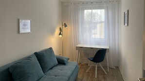 Pieni, viihtyisä huone, jossa on harmaa sohva, valkoinen työpöytä, sininen tuoli ja suuri ikkuna, jossa on läpikuultavat verhot.