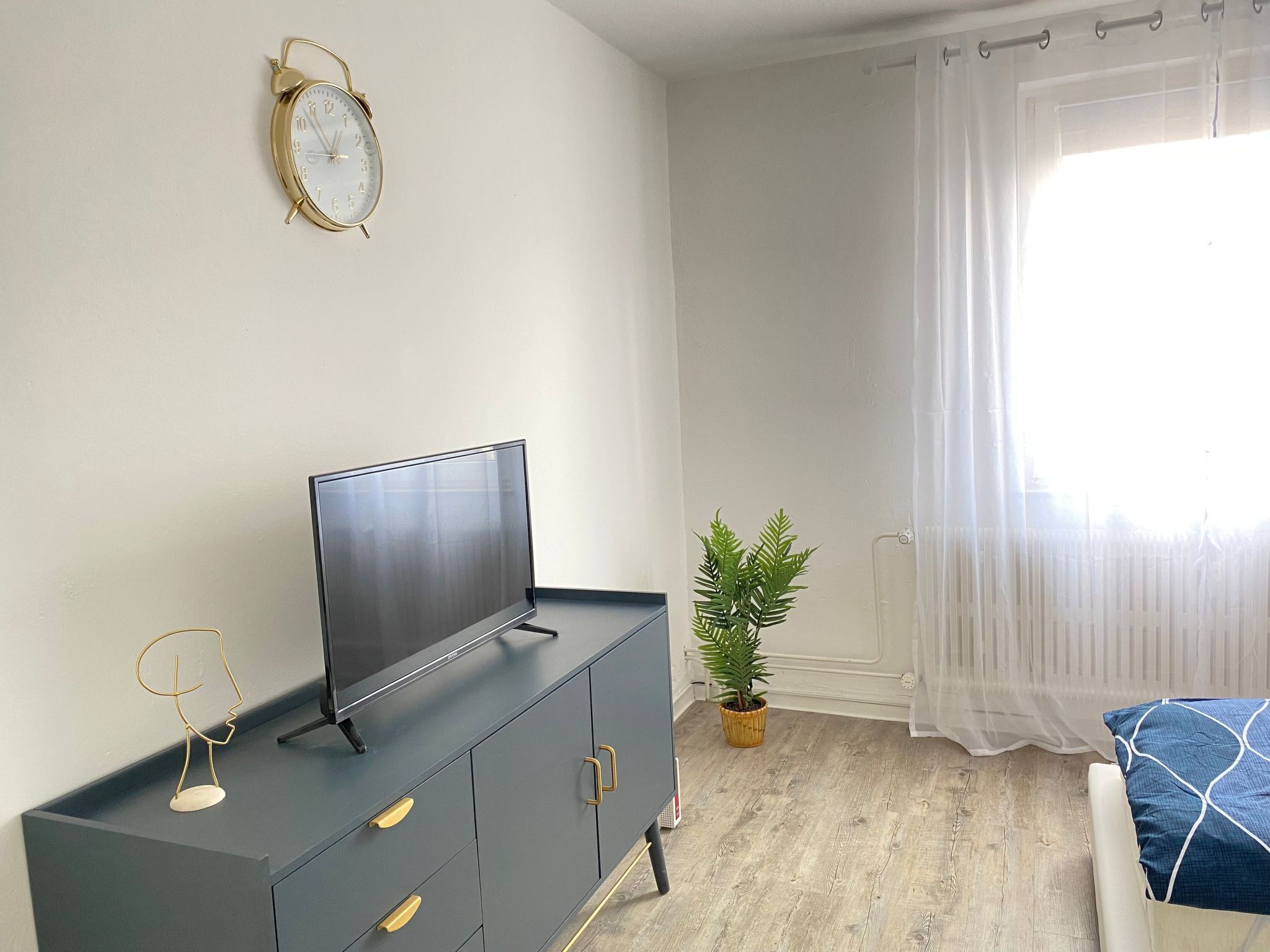 Ein Zimmer mit Fernseher auf einem dunklen Schrank, einer Wanduhr, einer Topfpflanze und einem Fenster mit transparenten Vorhängen.