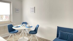 Moderni huone, jossa on pyöreä ruokapöytä, siniset tuolit, valkoinen sohva ja ikkuna, jossa on sälekaihtimet.