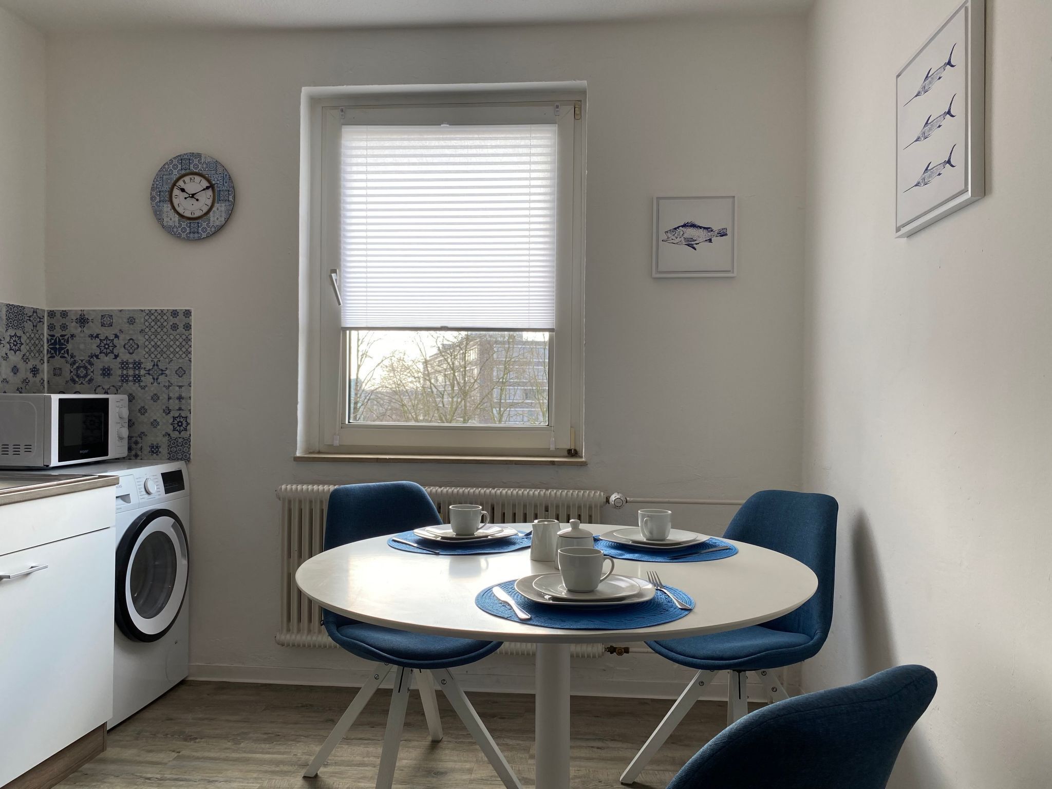 Een kleine keuken met een magnetron, wasmachine en een eettafel met blauwe stoelen.