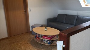 Ein kleiner, gemütlicher Raum mit Holztür, einem grauen Sofa, einem Tisch mit Lampe und einem Oberlicht.