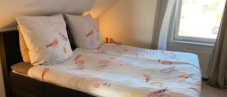 A bed with a patterned bedspread and two pillows, a window with curtains, and a radiator.