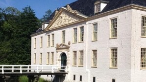 Ein großes, weißes Gebäude mit einer symmetrischen Fassade, einem zentralen Eingang mit dekorativem Bogen und einer Brücke über ruhigem Wasser.
