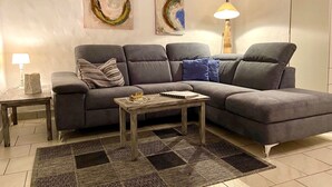 Ein modernes Wohnzimmer mit einem grauen Ecksofa, einem Holztisch und einem gemusterten Teppich. An den Wänden hängen abstrakte Gemälde und eine Stehlampe steht bereit.