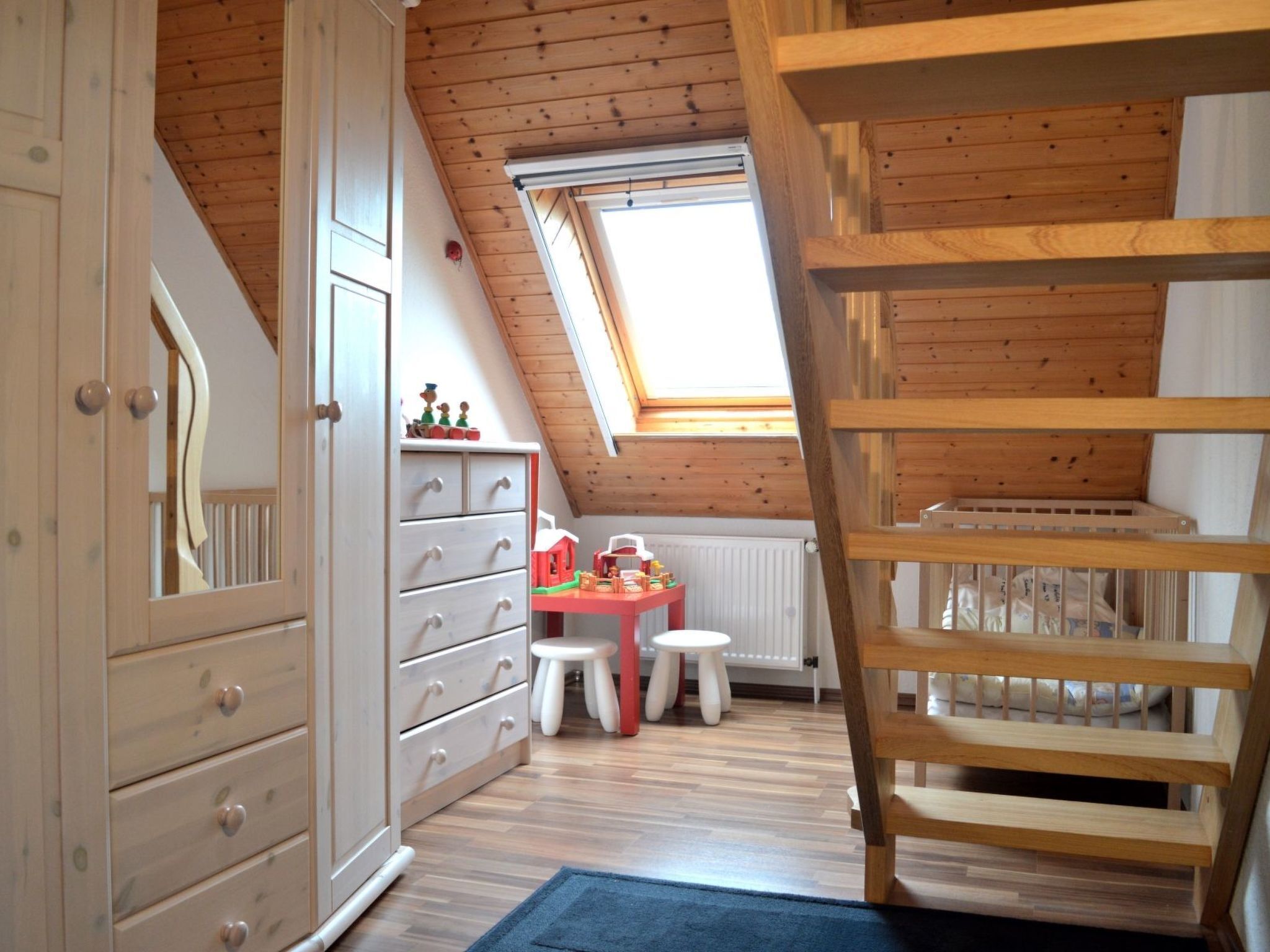 Une pièce avec un plafond en pente, des poutres en bois, un lit en mezzanine, une commode, une petite table avec des jouets et une fenêtre.
