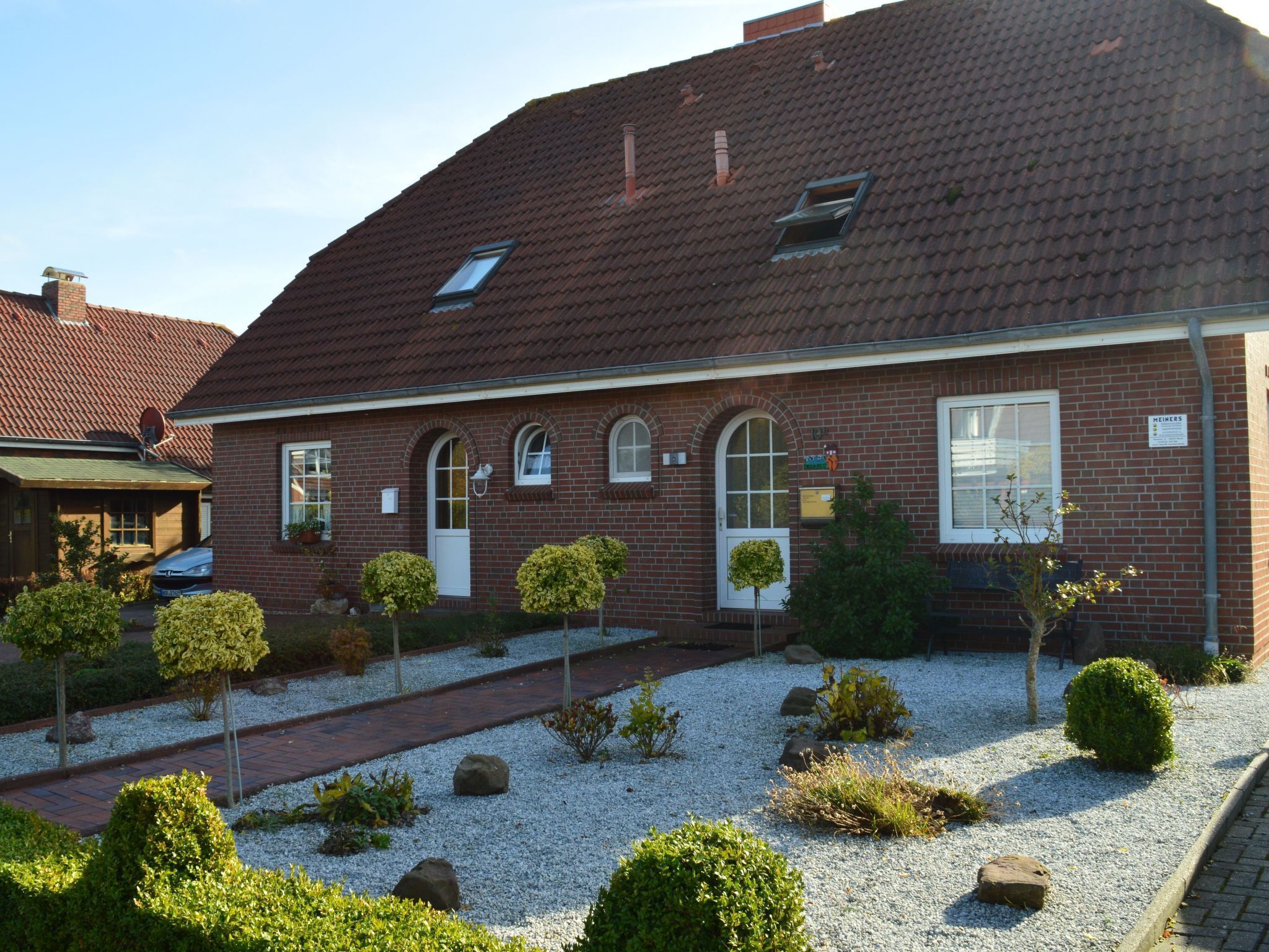 Een gebouw van baksteen met een schuin dak, een kleine tuin met grind en struiken, en een pad dat naar de ingang leidt.