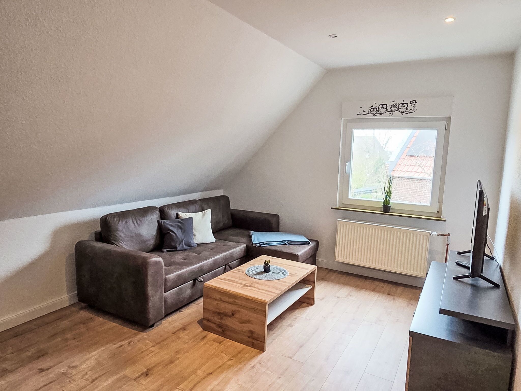 Ein modernes Wohnzimmer mit einem braunen Ledersofa, einem Holztisch, einer schwarzen TV-Konsole und einem Fenster mit Blick auf ein Gebäude.