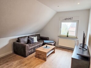 Ein modernes Wohnzimmer mit einem braunen Ledersofa, einem Holztisch, einer schwarzen TV-Konsole und einem Fenster mit Blick auf ein Gebäude.